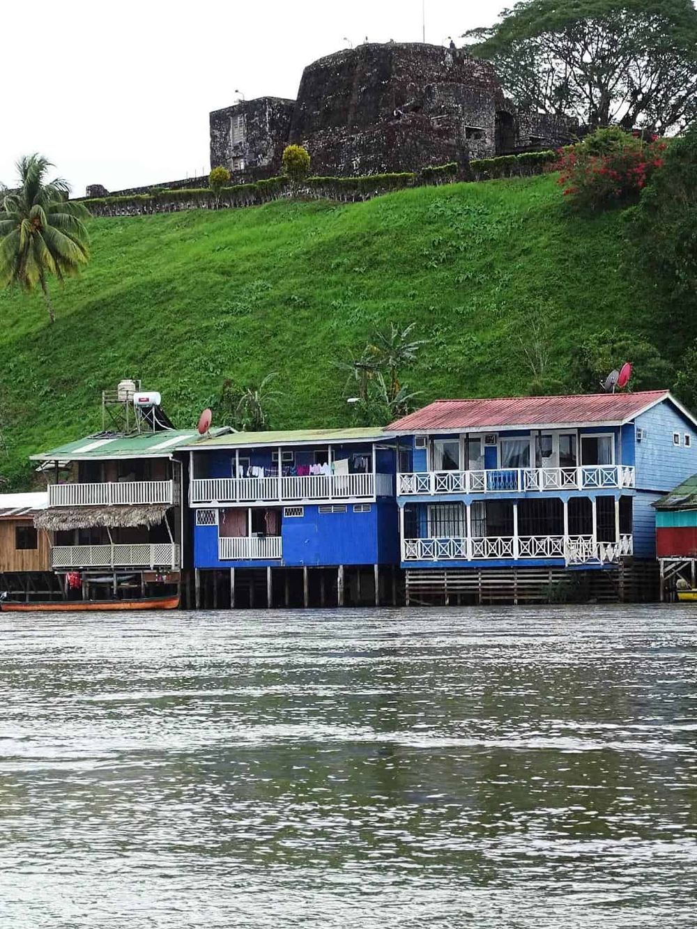Colorful waterfront houses in a tropical setting with lush green hills and historical ruins in the background.