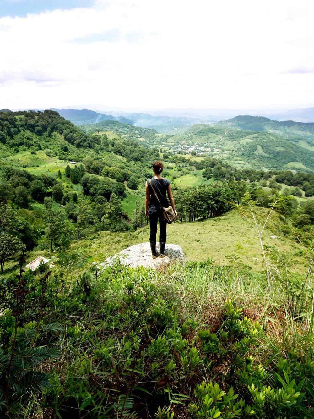 Vast green valley view with woman standing on rock looking at scenic landscape, hills, and lush nature.