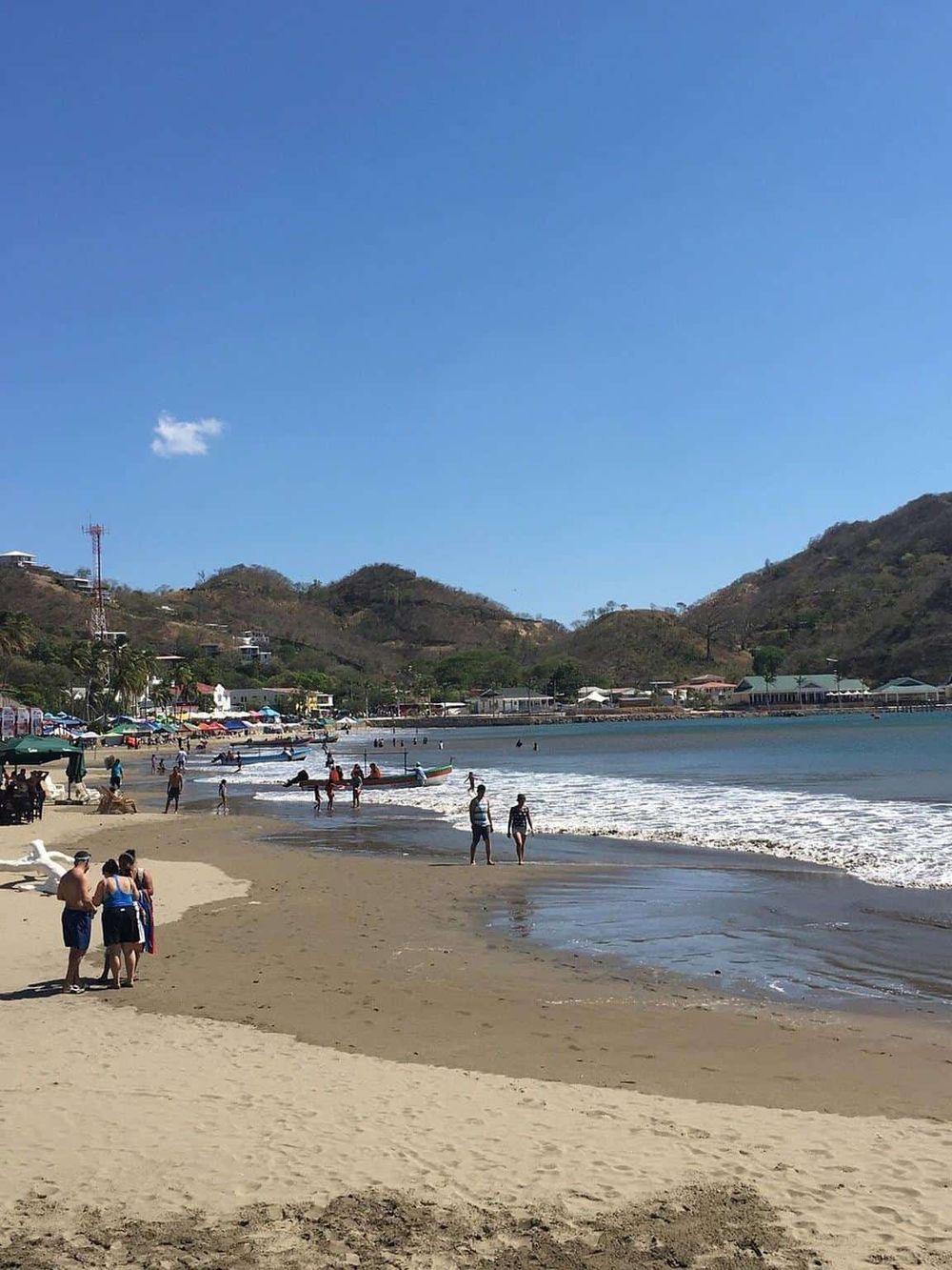 Scenic beach with blue sky, hills, and people enjoying the coastal view and water activities.