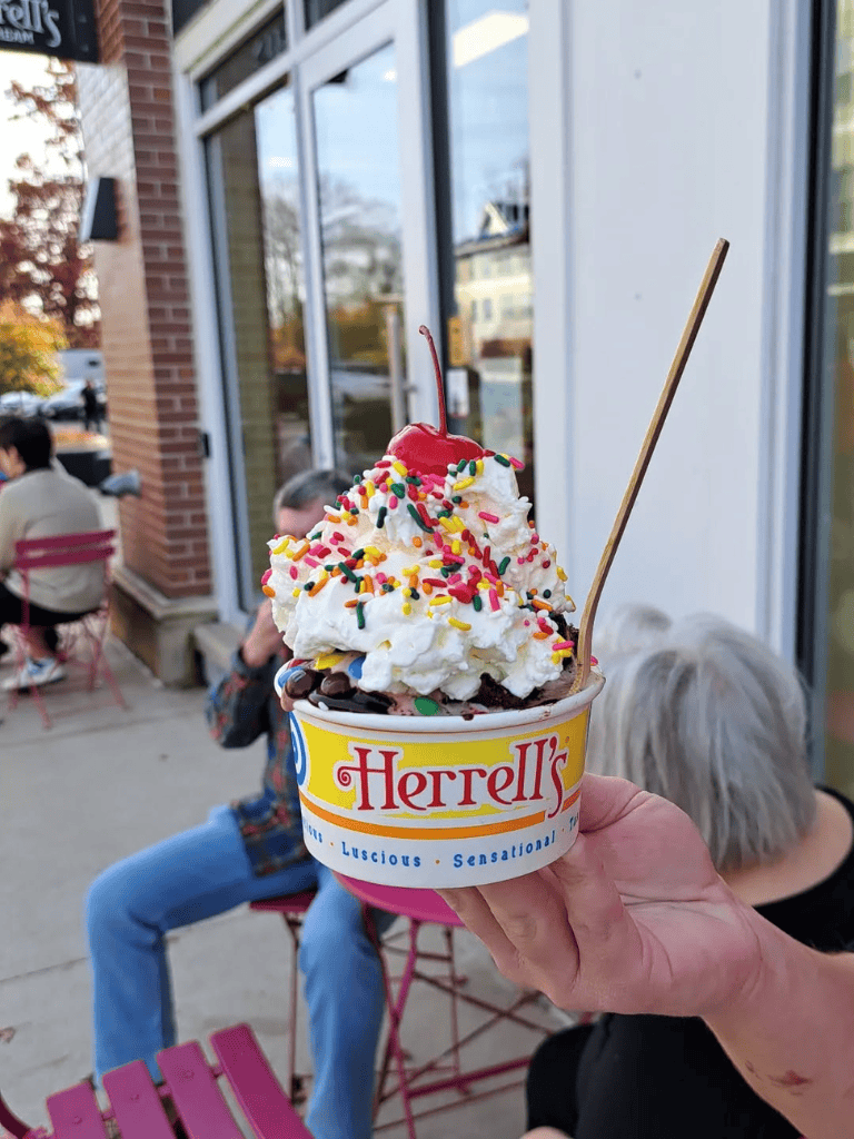 Creamy ice cream in Herrell’s cup with sprinkles, cherry, and whipped cream, outdoor seating scene.