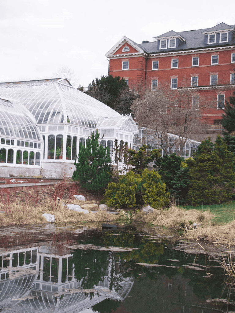 Greenhouse with a historic building in the background, lush landscape, and pond in foreground.