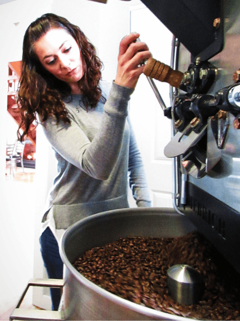 Freshly roasted coffee beans being poured from a commercial coffee roaster by a woman in a modern kitchen.