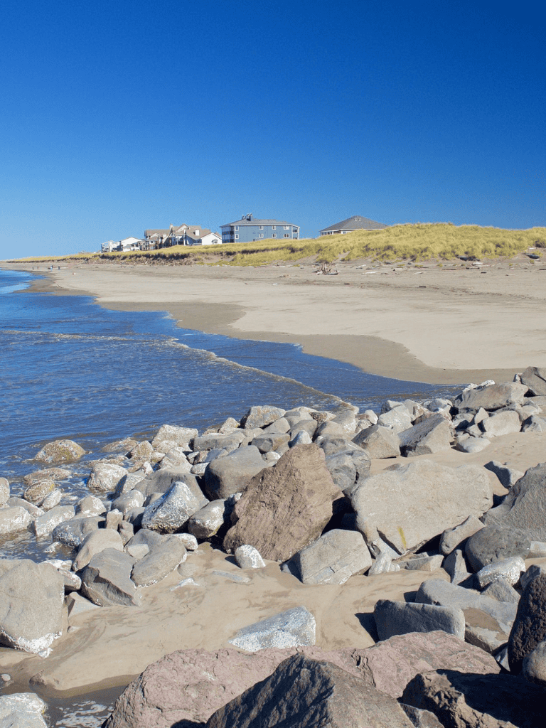 Seaside beach with rocks, sand dunes, and houses under a clear blue sky.