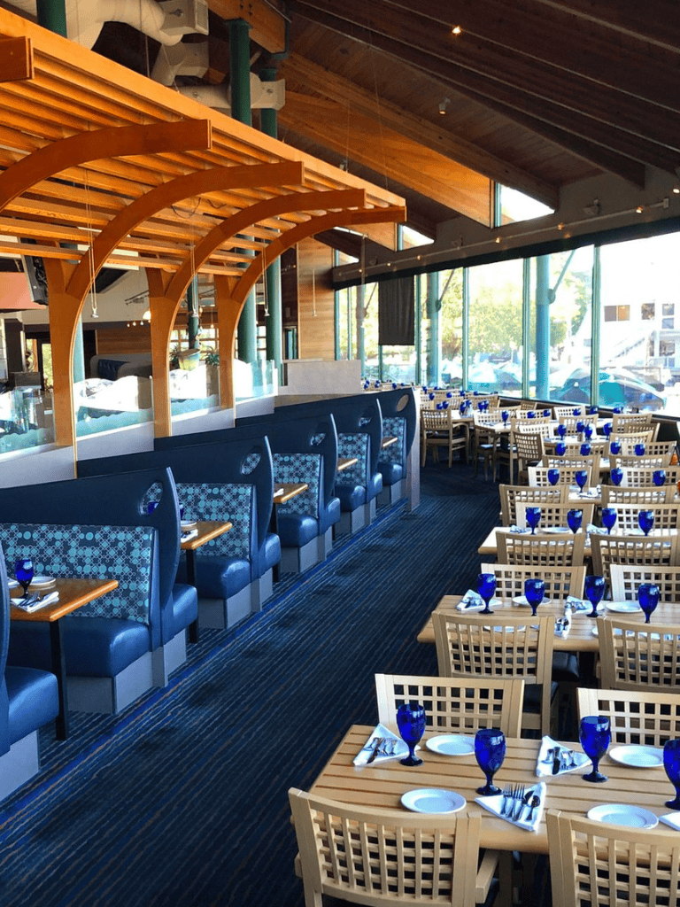 Elegant restaurant interior with wooden accents and large windows, set for dining with blue glassware and table settings.
