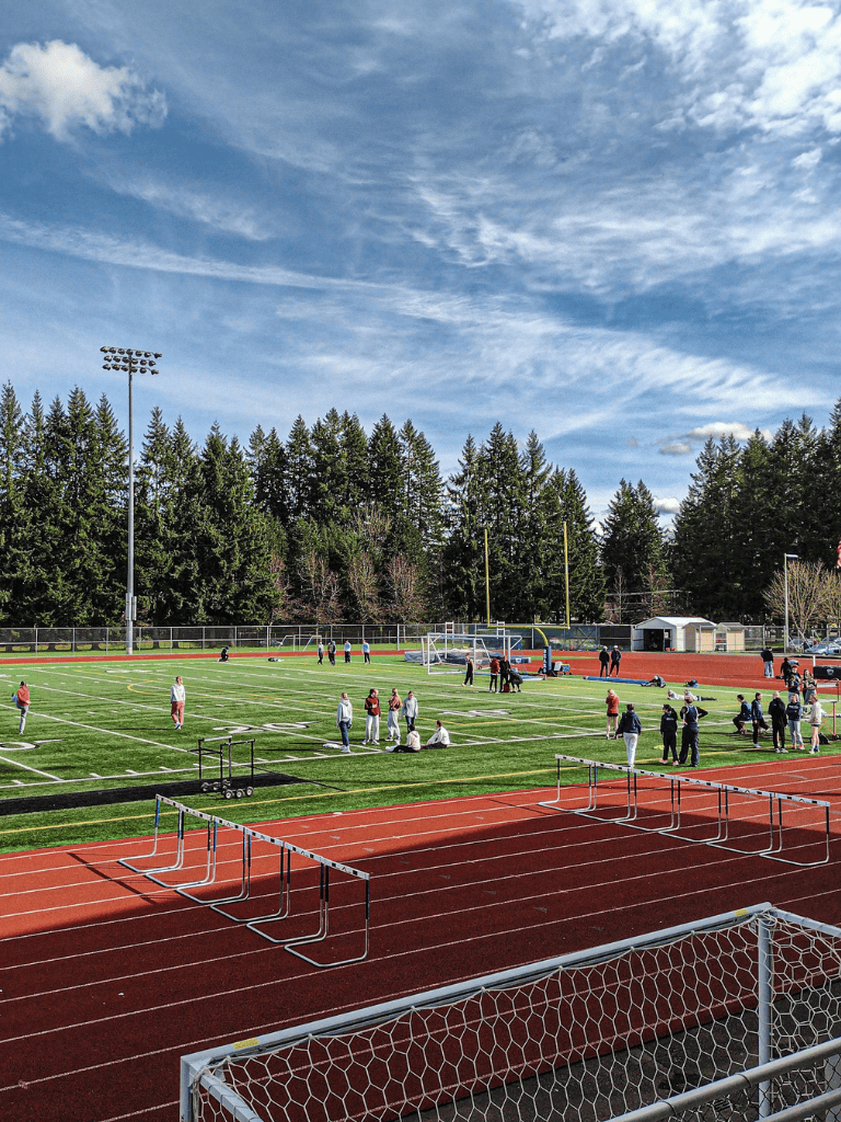 High school football practice and track field with hurdles in the foreground, sports field, and trees under a blue sky.