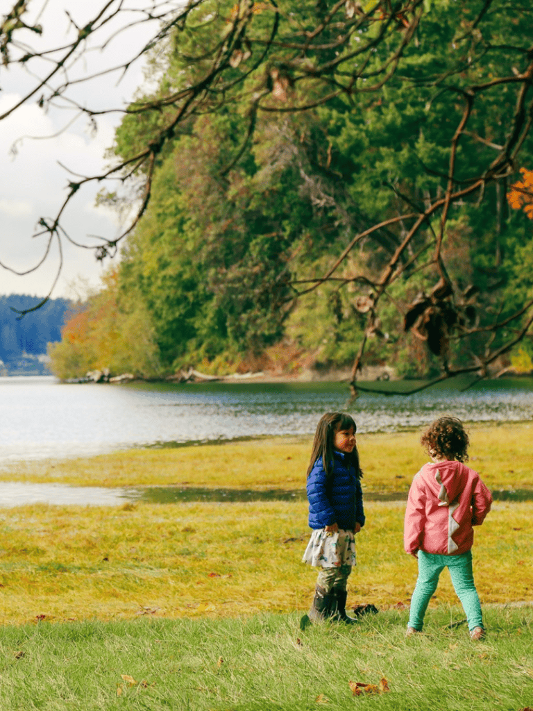 Children playing near a river in a lush, scenic outdoor park with autumn foliage.