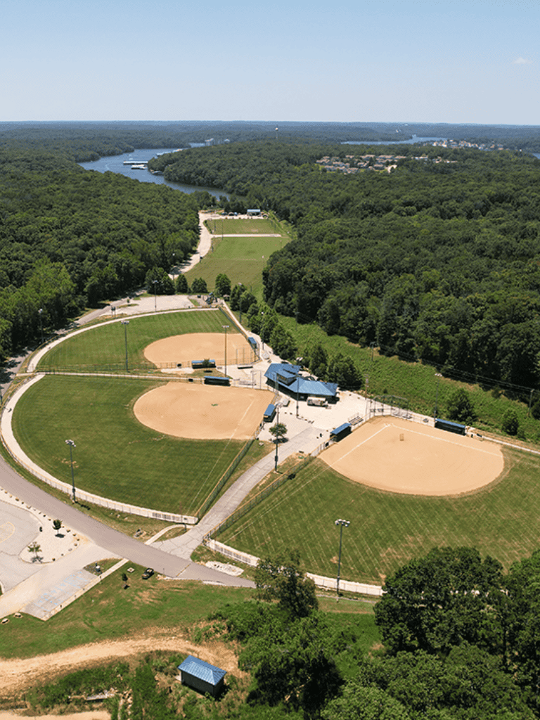 Ballfield complex with baseball diamonds and surrounding greenery near a river and wooded area.