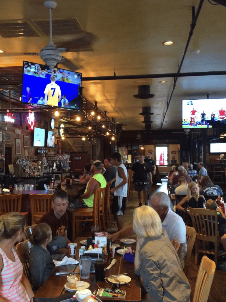 Large sports bar with multiple TV screens showing live sports, crowded with diverse patrons enjoying food and drinks.