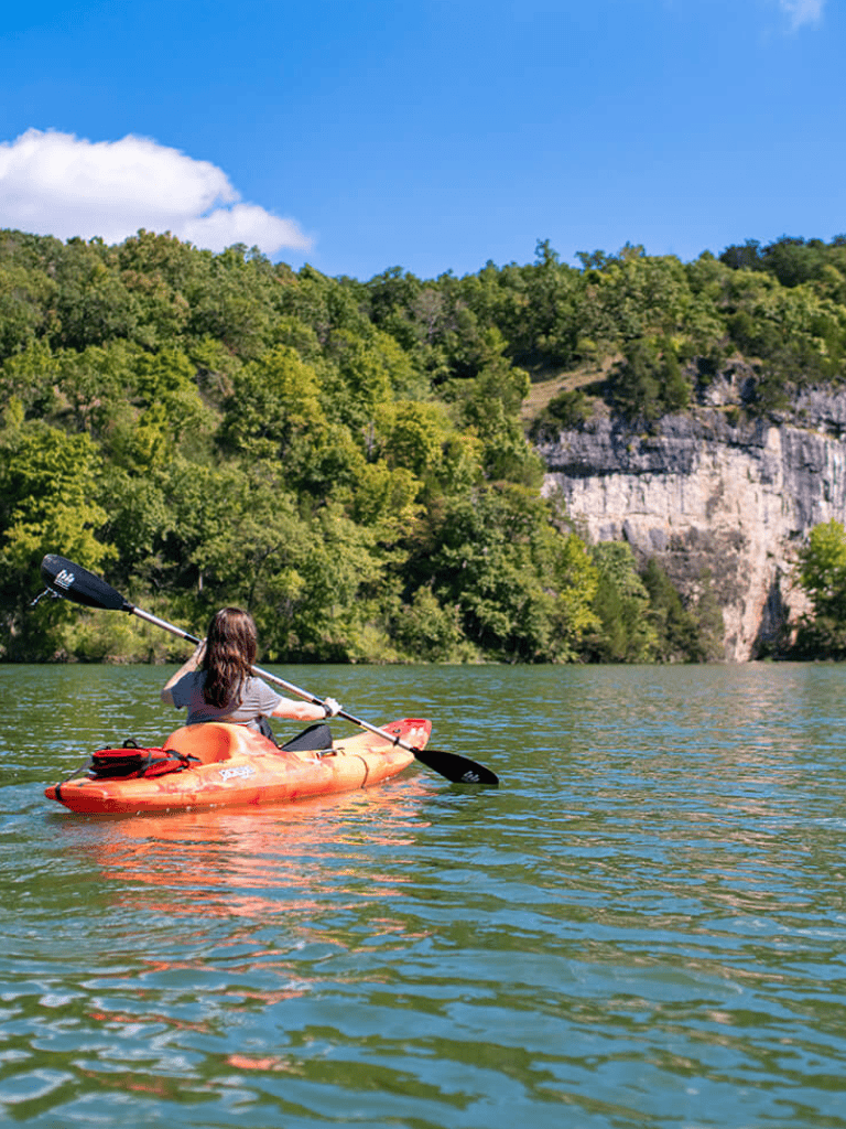 Kayaking on scenic river with lush green trees and rocky cliffs in the background.
