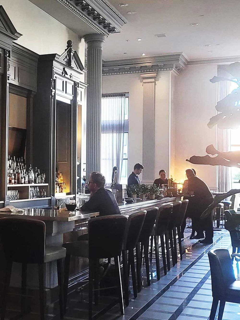 Modern hotel bar with upscale decor, high-back chairs, and bartenders serving drinks.