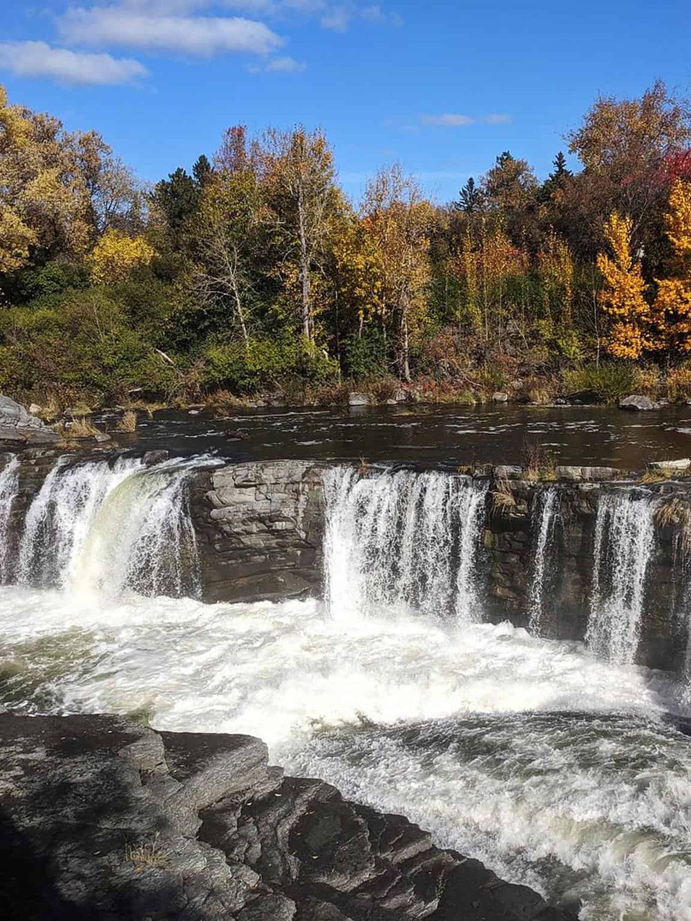 Colorful autumn trees along a flowing river with a small waterfall, showcasing scenic nature and outdoor adventure.