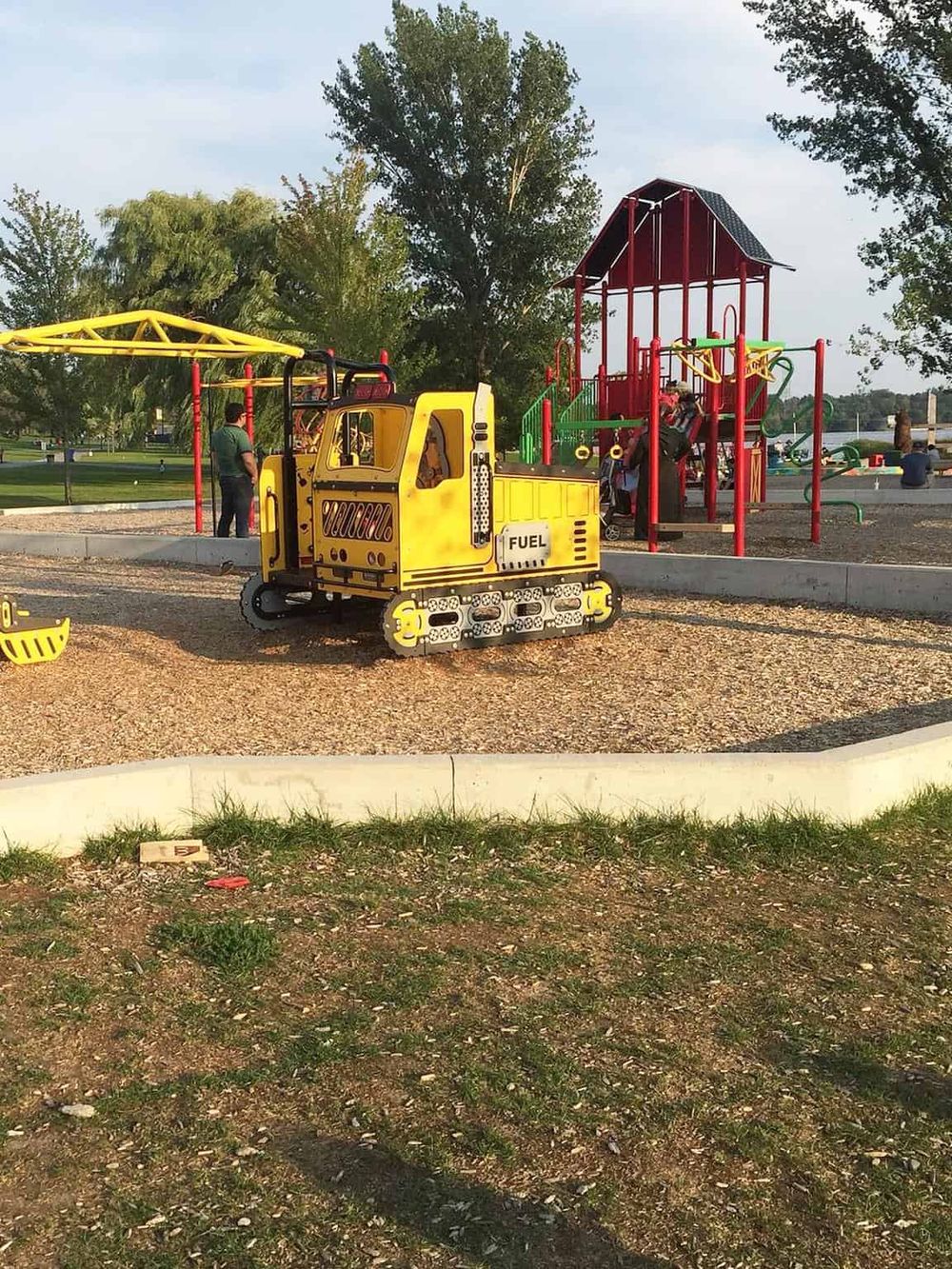 Colorful playground with a yellow construction-themed playset and red climbing structure in a park, perfect for kids' outdoor fun.