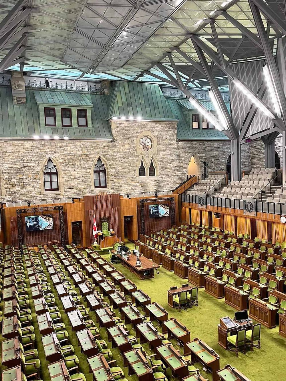 Modern legislative chamber with green chairs and wood accents, featuring a stone wall, high ceiling, and large screens for government meetings.