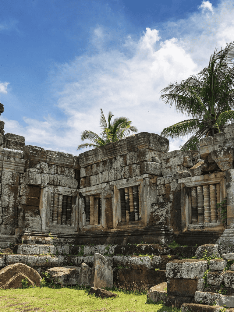 Ancient stone ruins with carved windows and tropical palm trees under a bright blue sky.