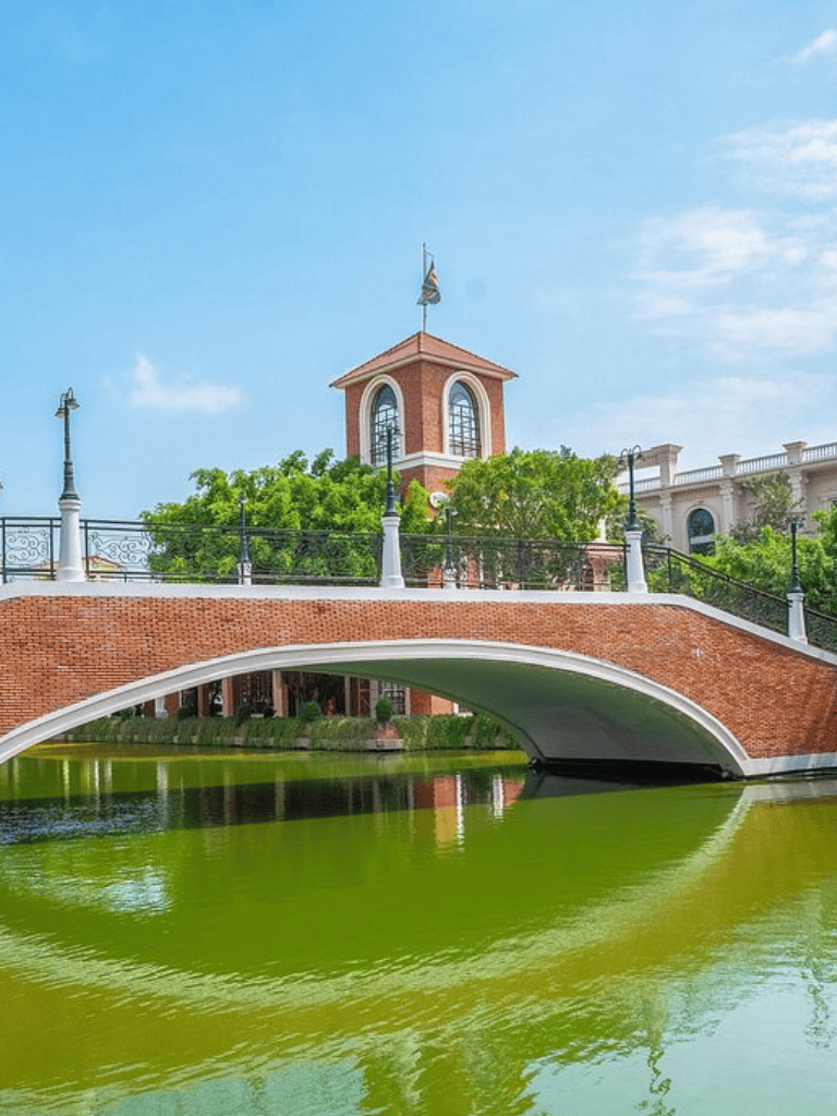 Charming brick bridge over a green pond with a red brick clock tower in the background.