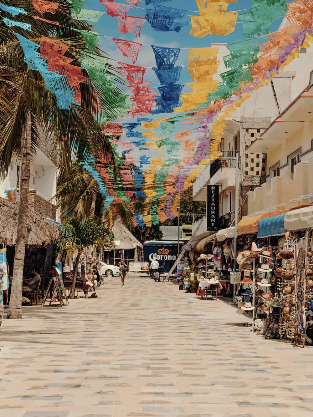 Colorful street with hanging papel picado banners, shops, and palm trees in a lively Mexican market.