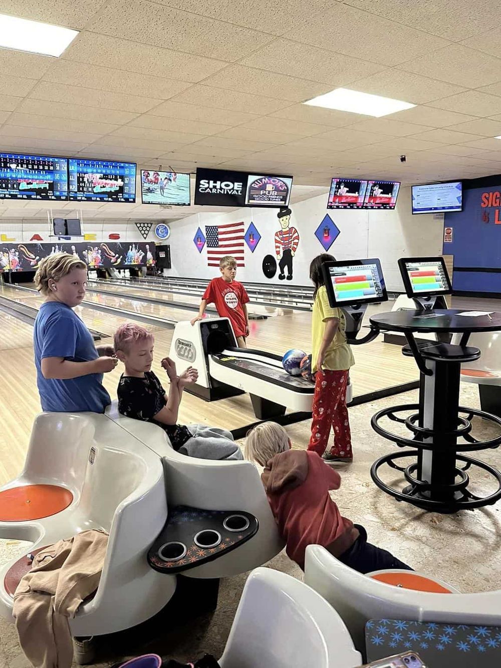 Bright indoor bowling alley with children, bowling balls, and electronic scoring screens.