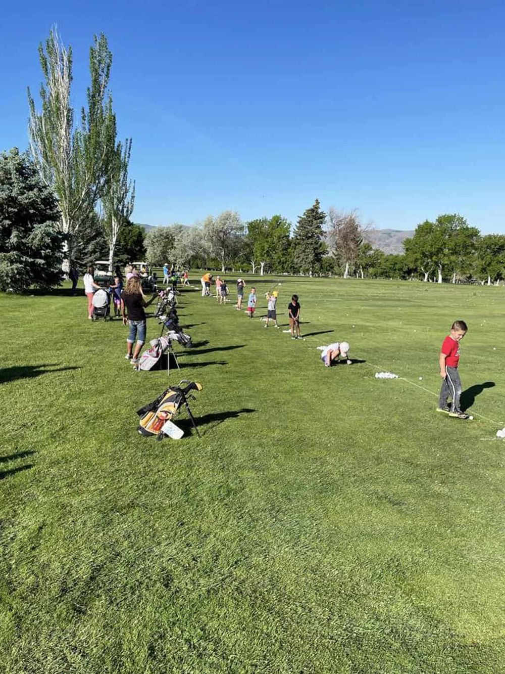 Children learning golf at a sunny outdoor golf course, with instructors and parents nearby.