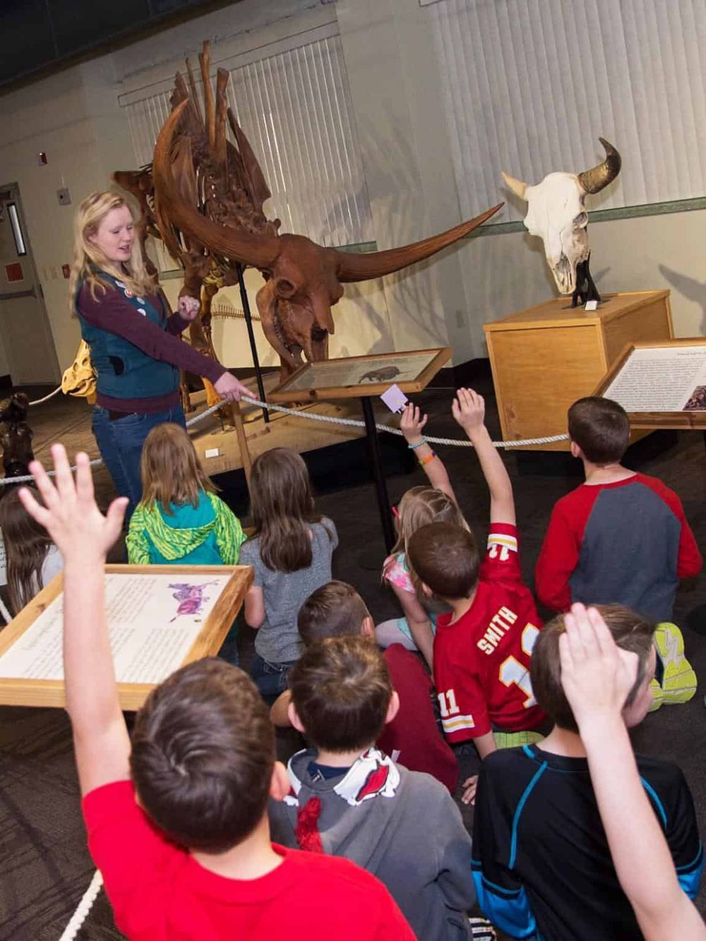 Colorful group of children engages with museum guide near dinosaur skeleton exhibit, educational field trip activity.