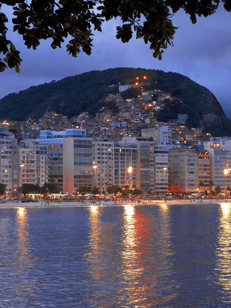 Vibrant cityscape of a coastal city at dusk with high-rise buildings and a hillside neighborhood illuminated.