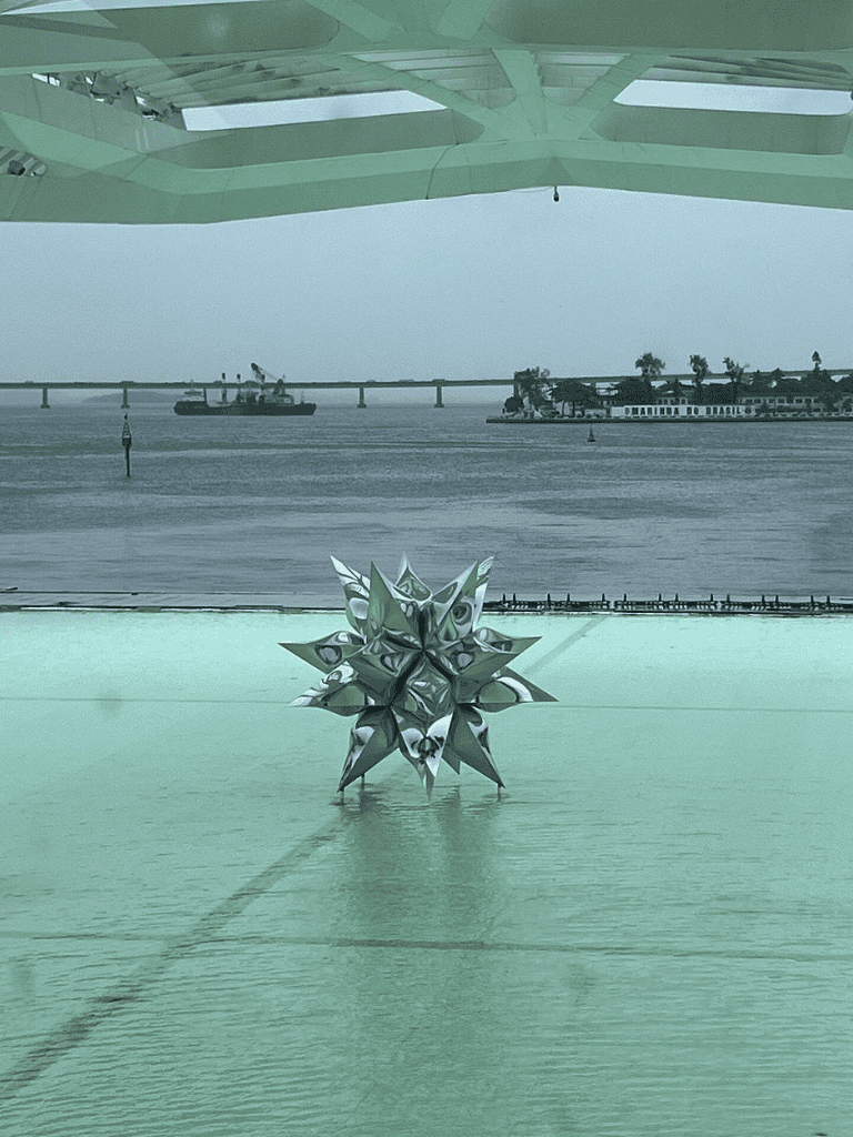 Shiny metallic geometric star sculpture on balcony overlooking river with bridge and boats.