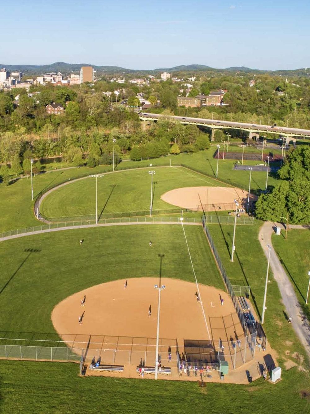 Aerial view of a baseball and softball park with green fields, surrounding trees, and cityscape in the background.