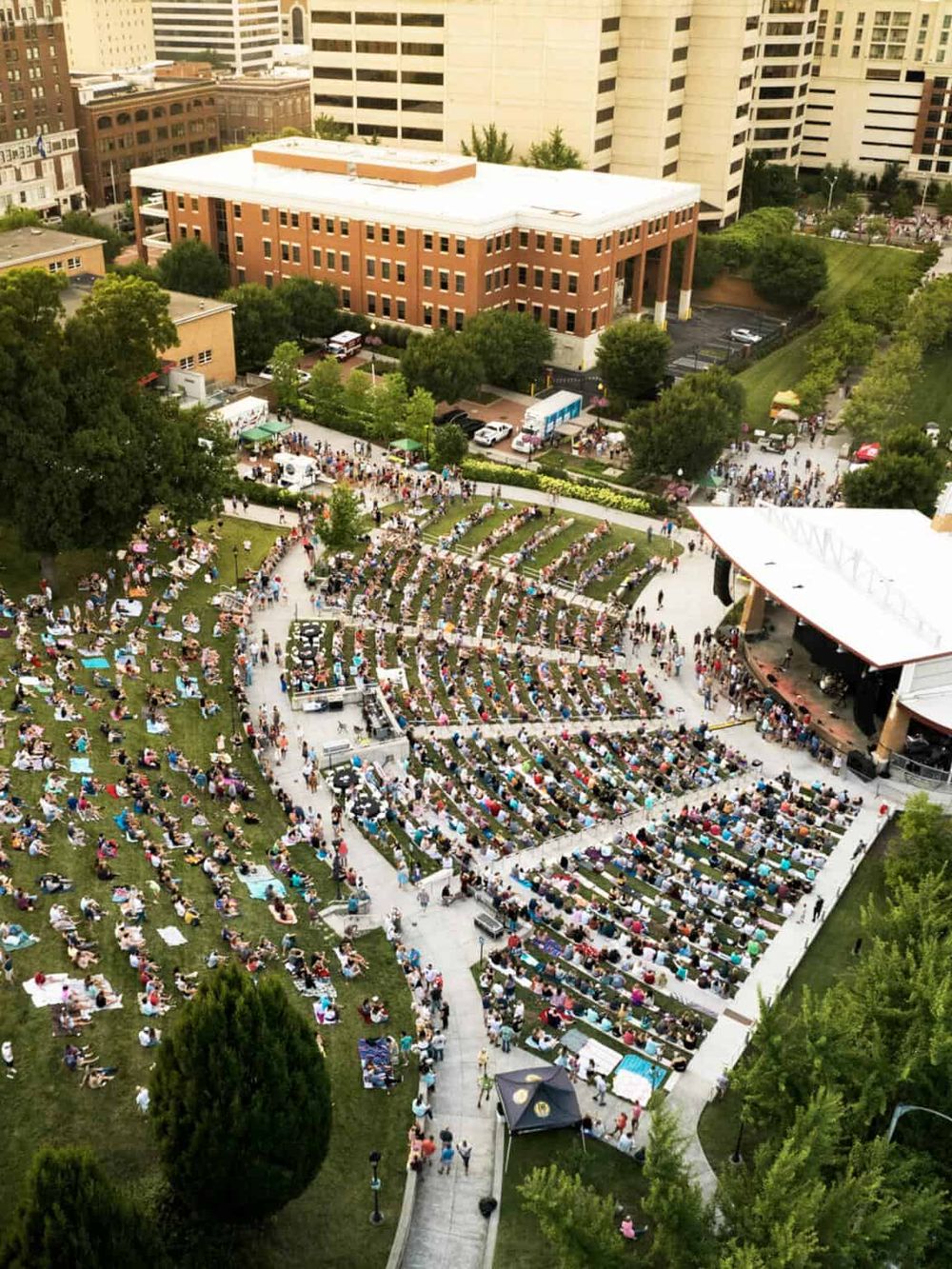 Crowd gathered at outdoor concert in urban park with city buildings in background.