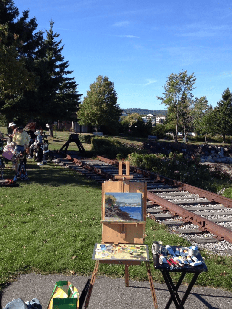 Painted landscape of a coastal scene near train tracks, outdoor art exhibit, vibrant green trees and clear blue sky.