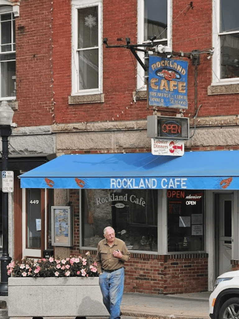 Cozy Rockland Cafe storefront with outdoor seating and a smiling man walking by, in a charming downtown setting.