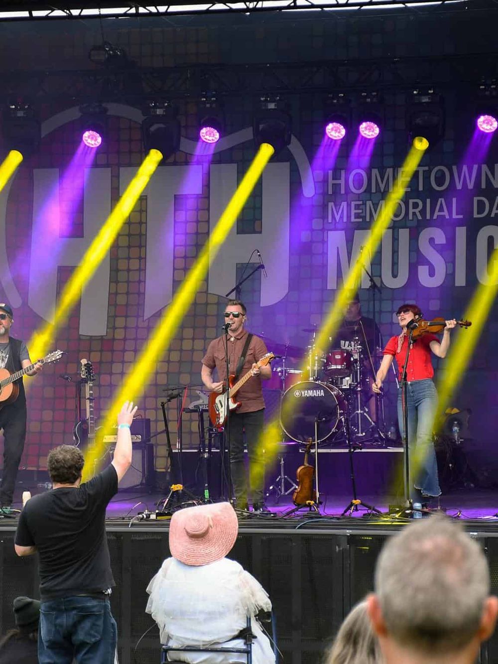 Live outdoor music performance with musicians playing guitar, violin, and drums on stage with colorful lighting.