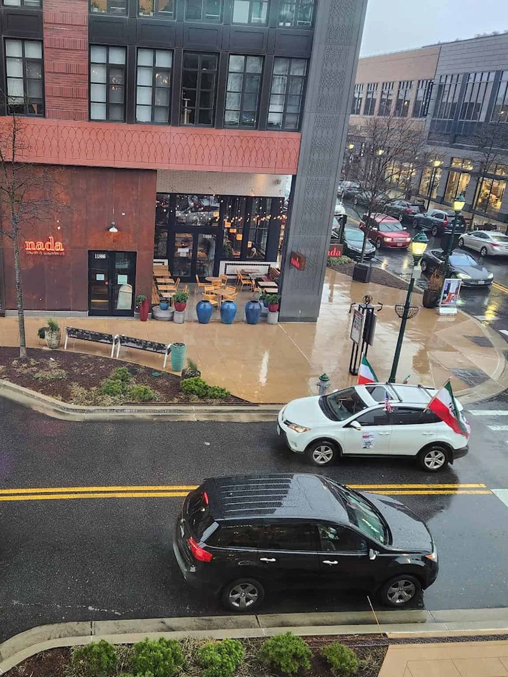 Modern urban street scene with parked cars, outdoor seating, and storefronts in a downtown area.