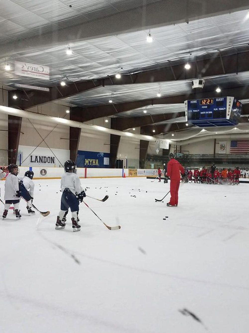Indoor ice skating rink with children practicing hockey and a coach, bright lighting, and scoreboard display.
