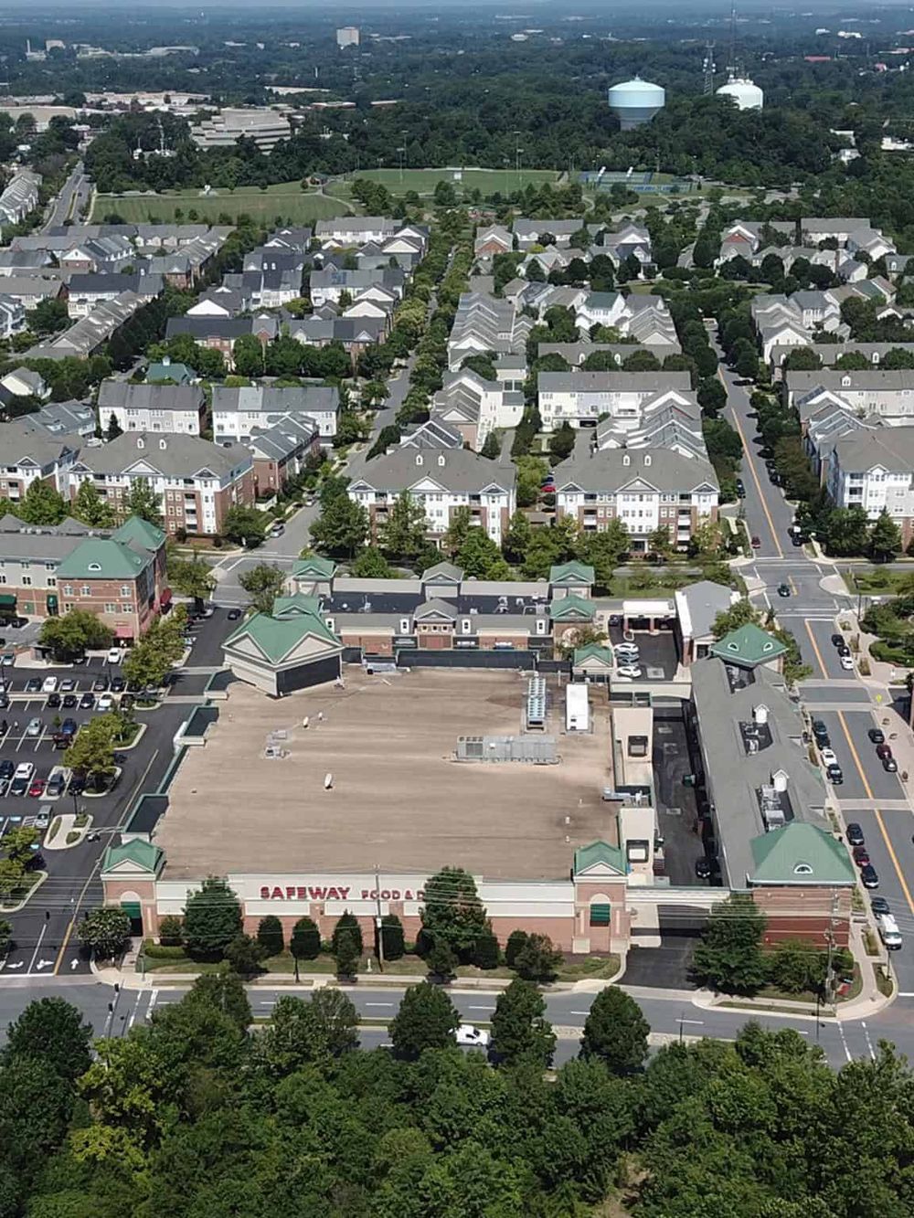 Empty shopping mall rooftop parking lot during daytime, surrounding residential neighborhood, greenery, and water towers in background.