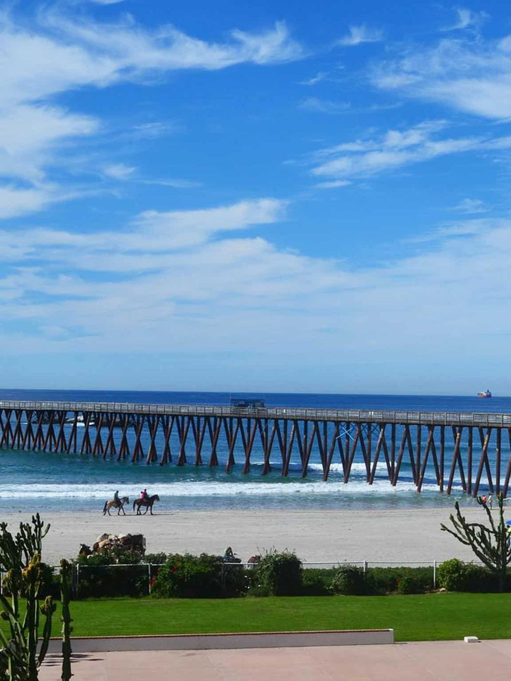 Coastal pier with horses riding near the beach and boats in the distance under blue sky.