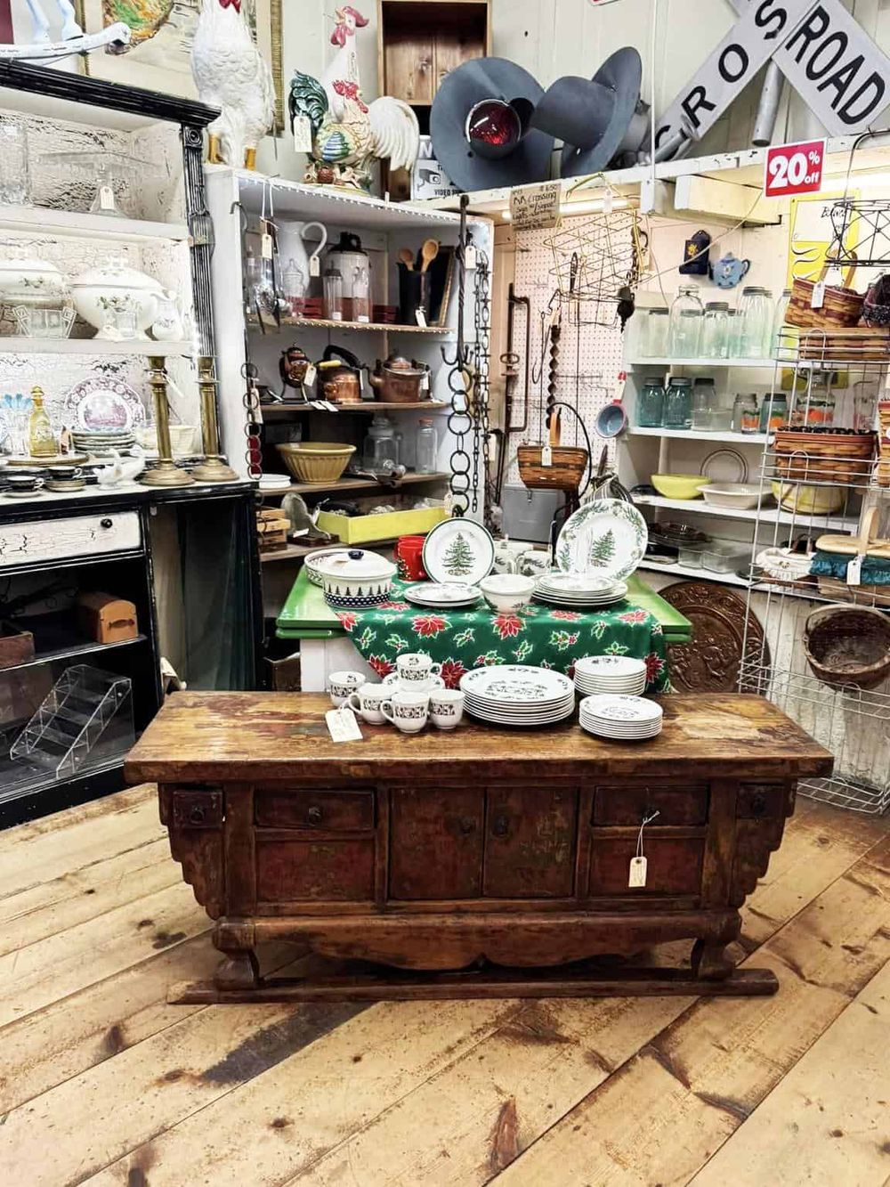 Vintage antique store with rustic wooden table displaying china plates and cups, surrounded by shelves of collectibles and décor.