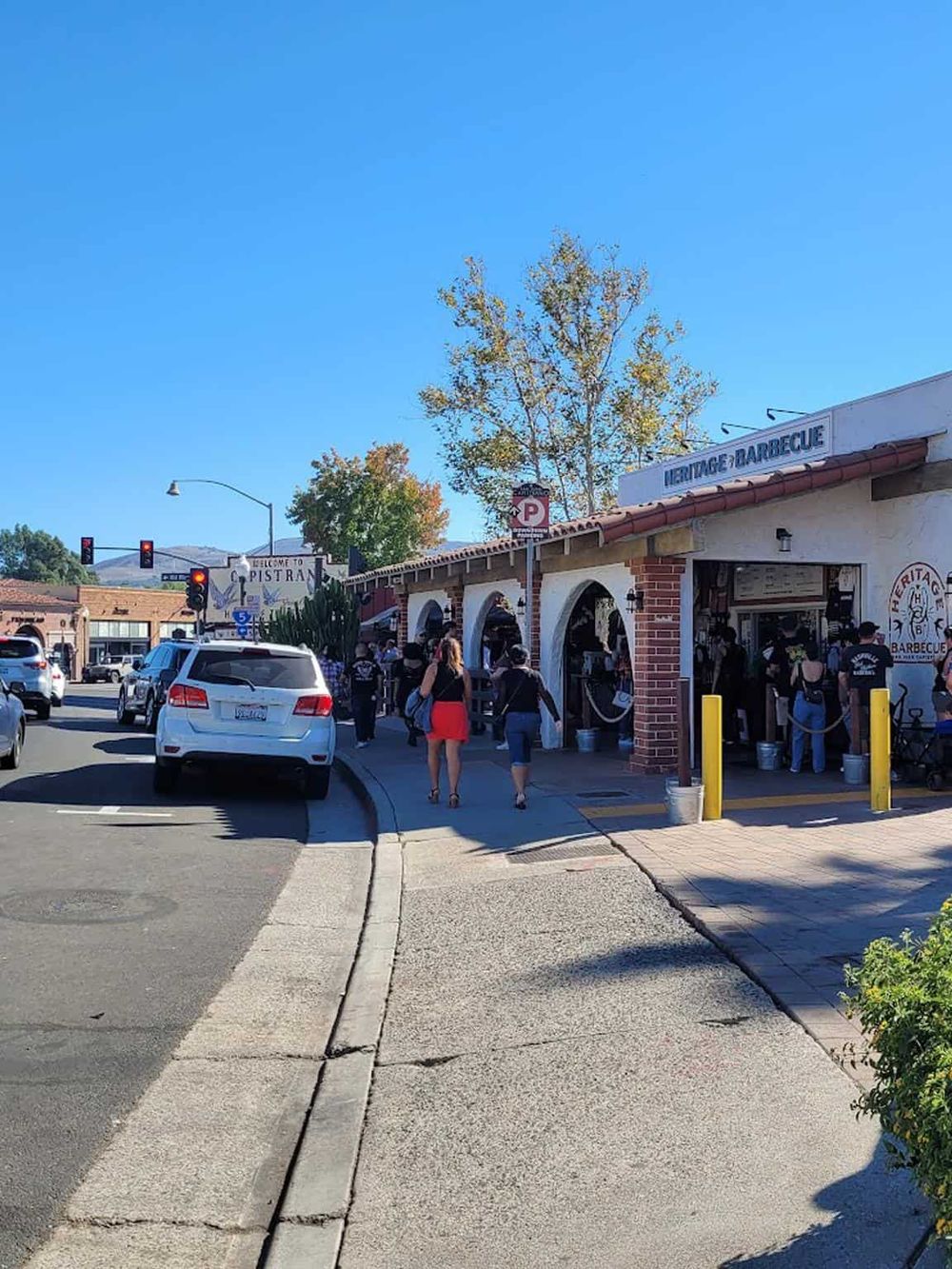 Outdoor street scene at a heritage and barbecue restaurant with people walking and cars parked, sunny day in a lively town area.