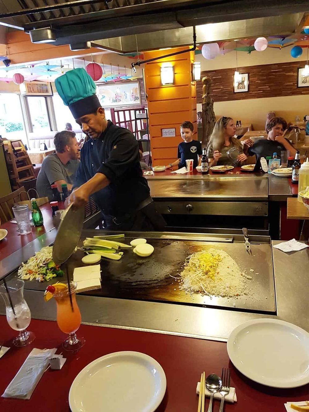 Vibrant teppanyaki restaurant with chef cooking on a hot griddle, surrounded by diners enjoying Asian cuisine.