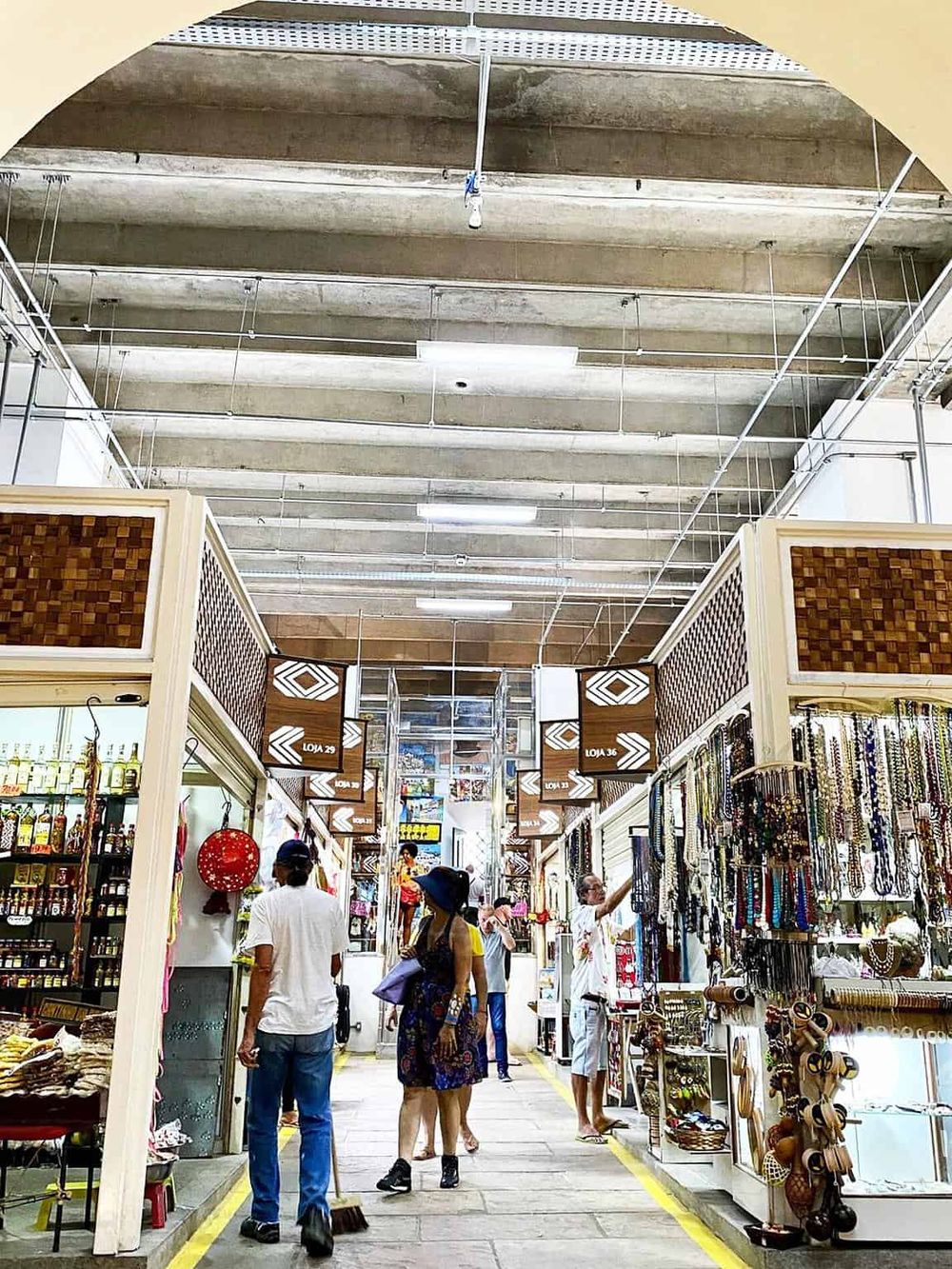 Colorful marketplace with shoppers browsing jewelry and souvenirs, under a high concrete ceiling.