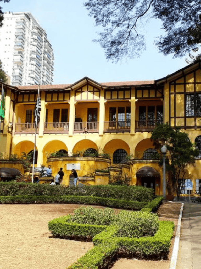 Historic yellow building with arches and balconies, surrounded by well-maintained gardens in an urban setting.