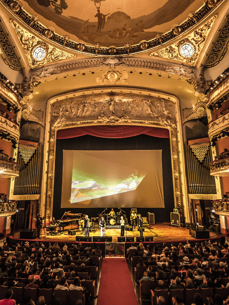 Elegant theater interior with a live music performance, ornate gold detailing, and an engaged audience.