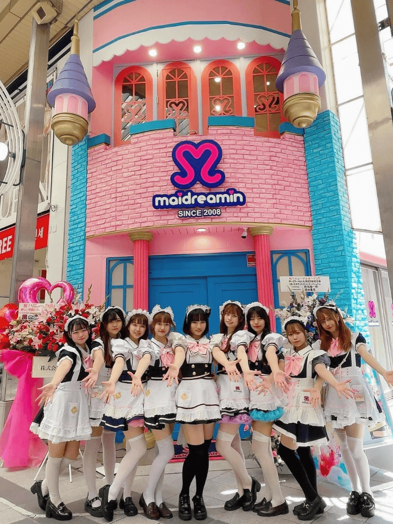 Pink castle-themed storefront with "maidreamin" logo, group of girls in maid costumes at Japan anime cafe.