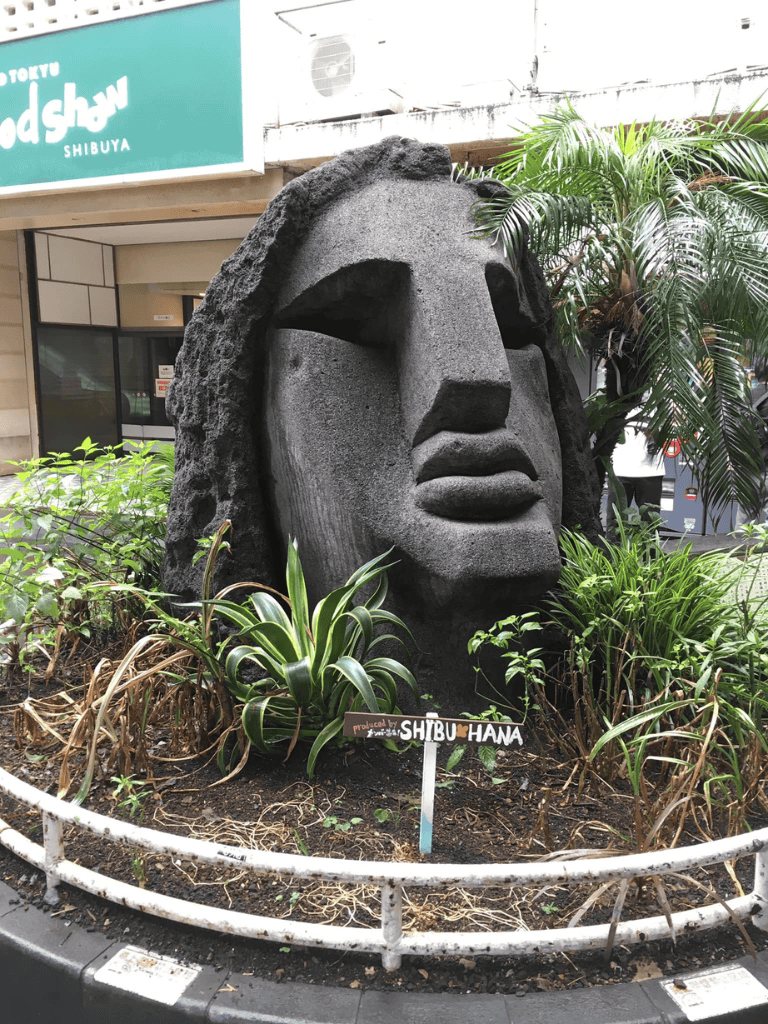 Stone sculpture of a human face from Shibuya, Japan, surrounded by lush plants.