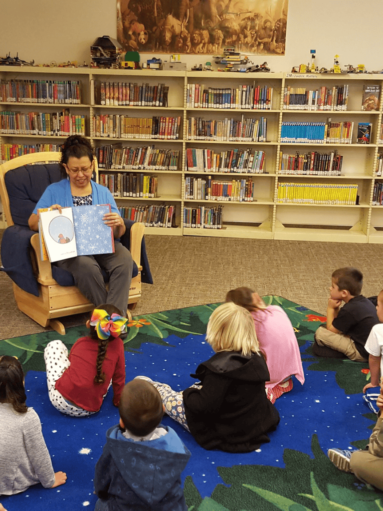 Childrens storytime at library with female librarian reading aloud to kids on colorful rug, shelves of books in background, engaging educational environment.