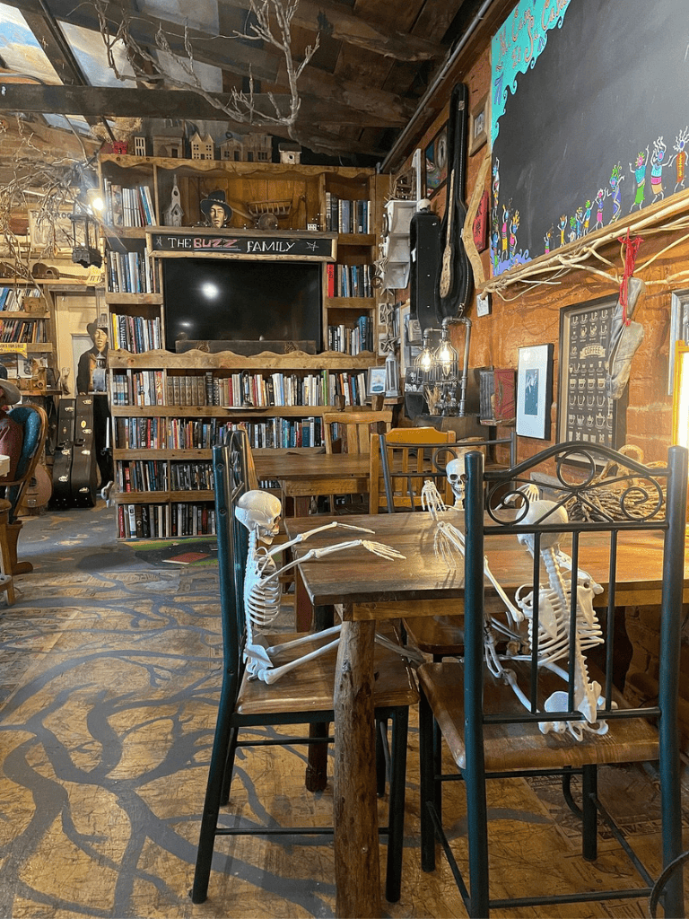 Skeletons sitting at a wooden table in a cozy, eclectic cafe filled with books and quirky decor.