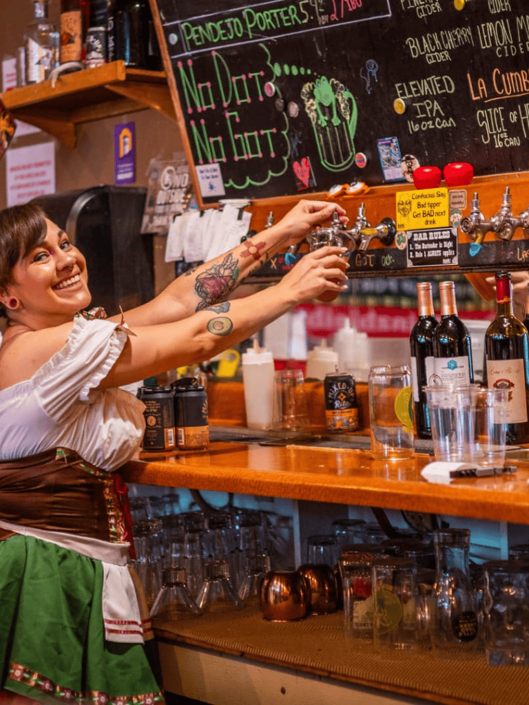 Cheerful bartender pouring beer at lively bar with colorful chalkboard menu and wine bottles, cozy pub ambiance.