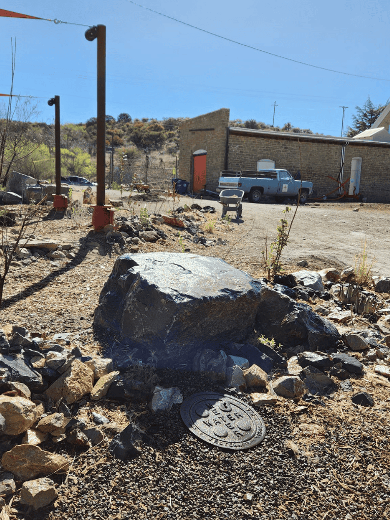 Weathered water meter box with large rock, under sunny sky at QuestForDirections location.