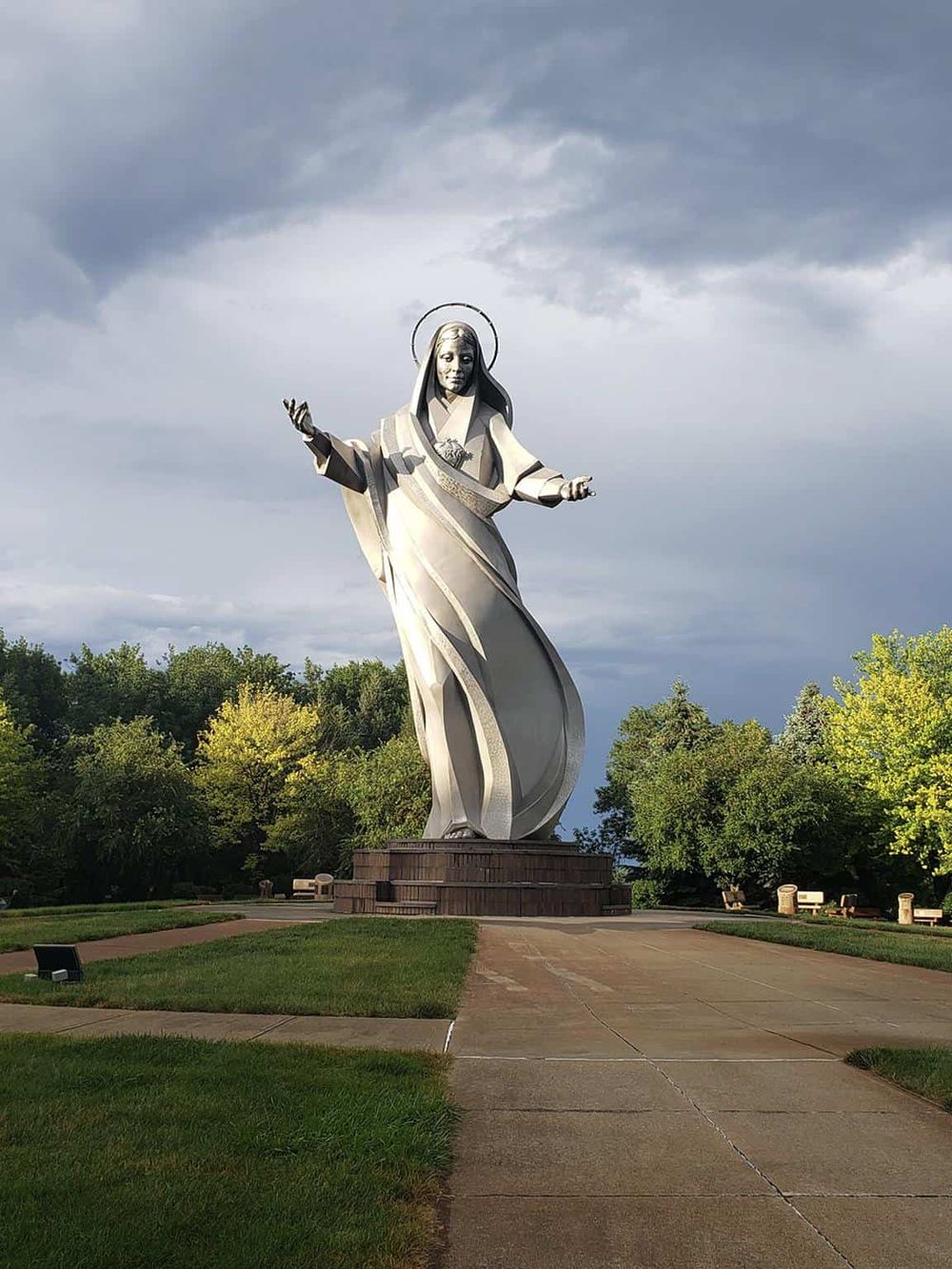 Majestic woman statue with open arms, symbolizing faith and guidance, set against a cloudy sky and surrounded by lush greenery.