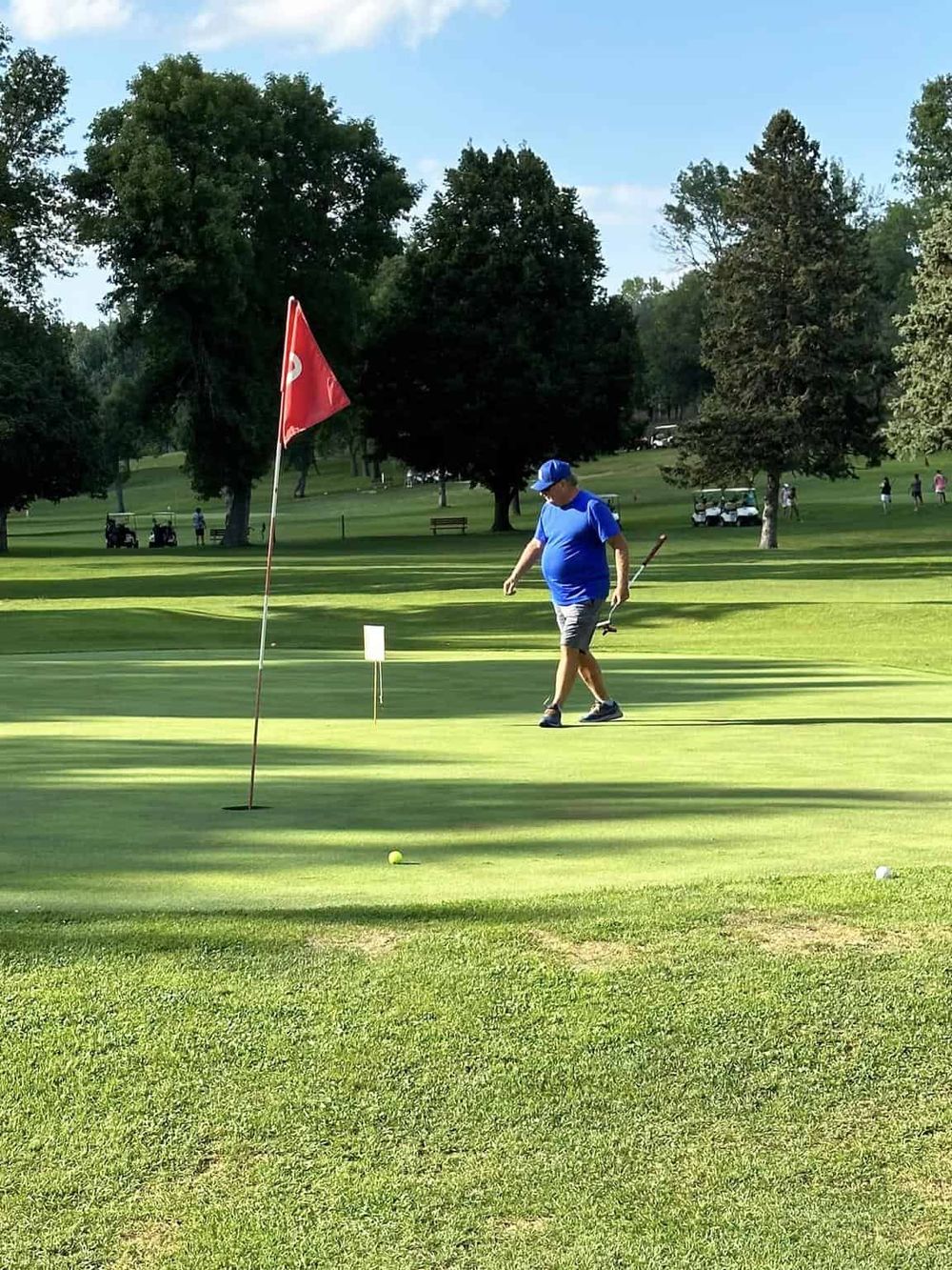 Golf course putting green with golfer preparing for a shot, scenic park setting, lush trees, and clear blue sky.