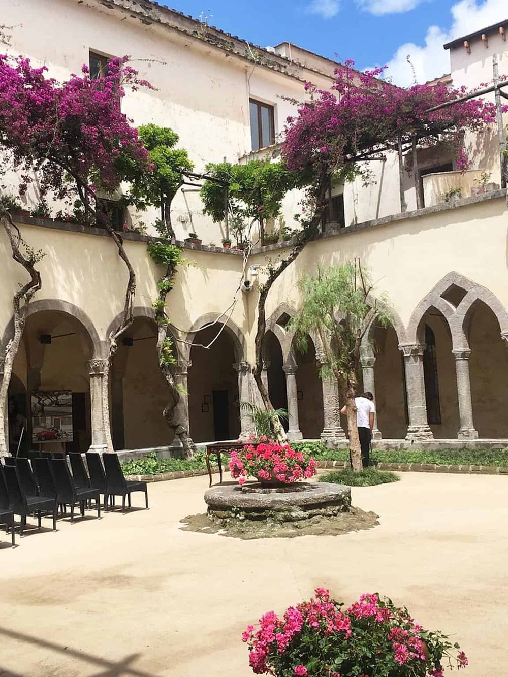 Picturesque courtyard with blooming pink flowers, old stone arches, and lush greenery in a historic European setting.