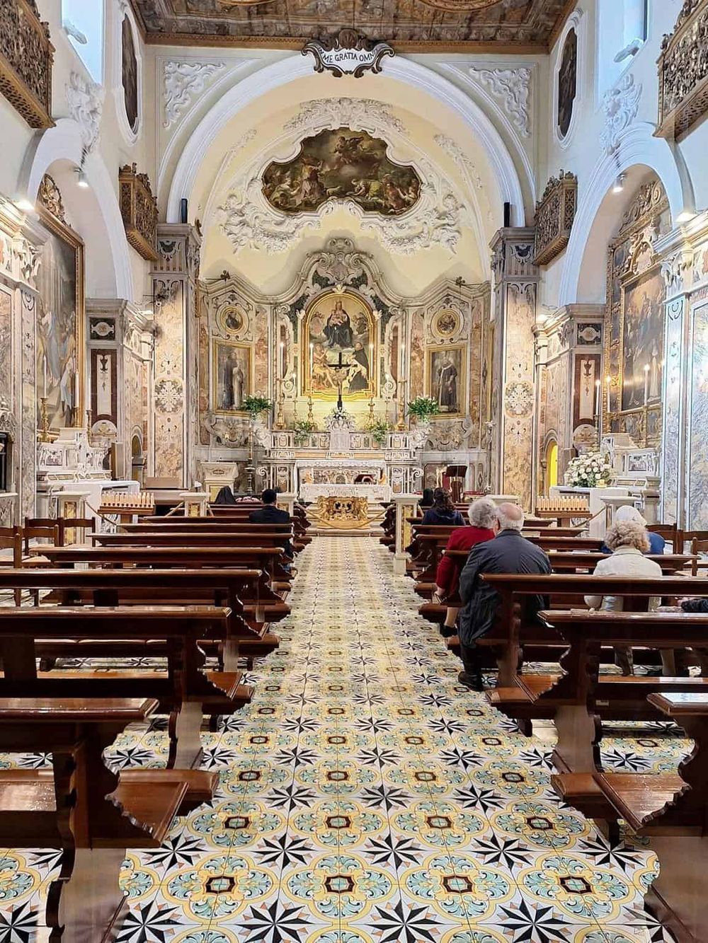 Intricate baroque church interior with ornate decorations, religious artworks, and worshippers seated in wooden pews.